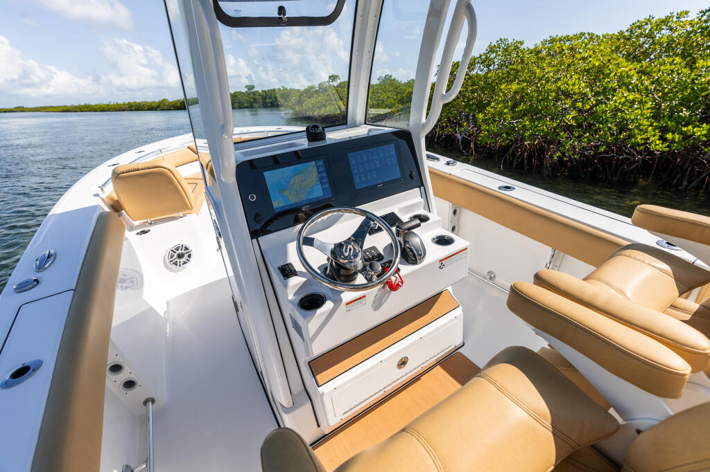 Helm station of a center console boat with dual GPS chartplotters, steering wheel, and tan upholstered seating in a mangrove-lined waterway
