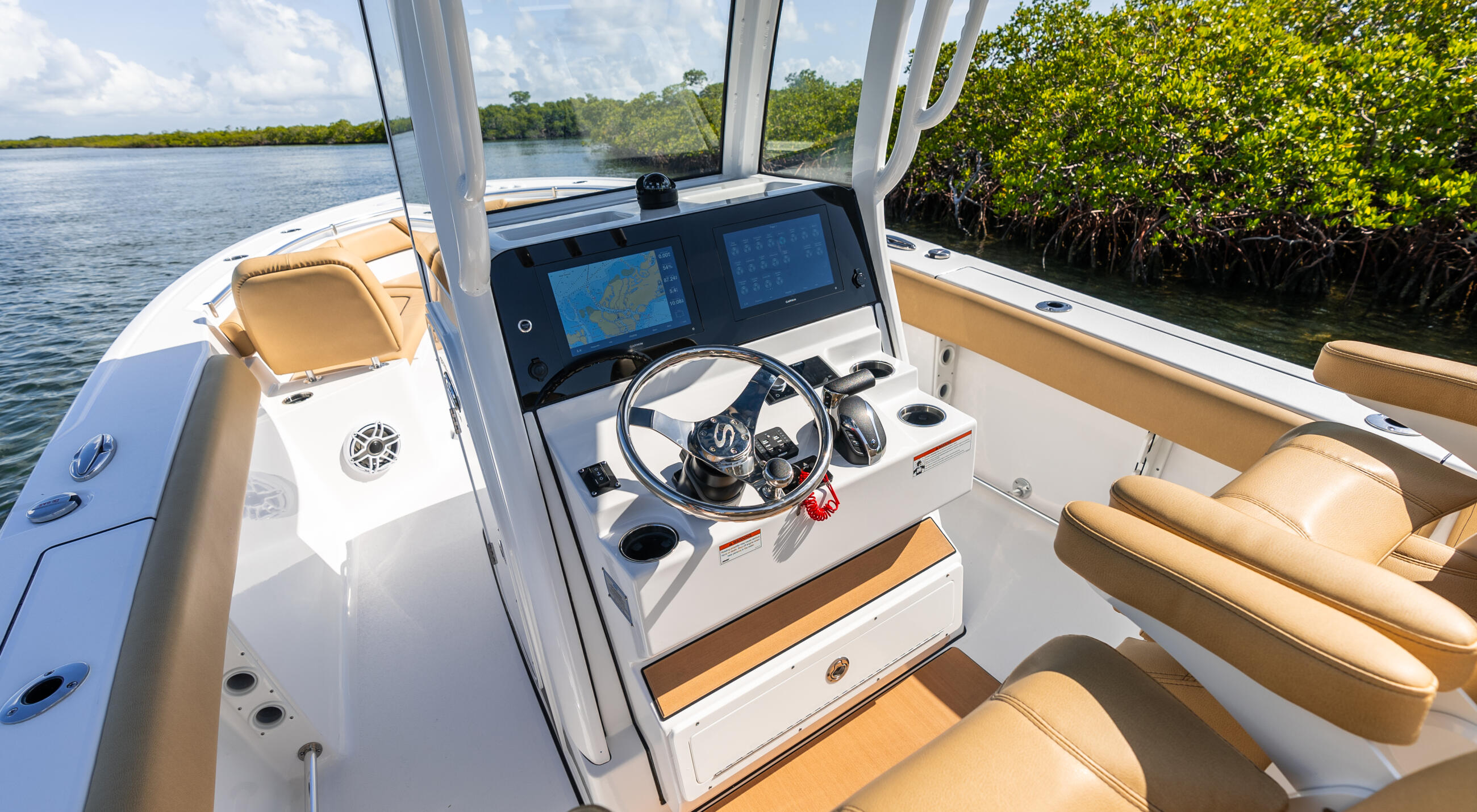 Helm station of a center console boat with dual GPS chartplotters, steering wheel, and tan upholstered seating in a mangrove-lined waterway