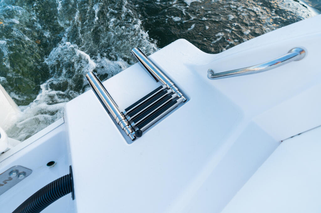 Overhead view of a white boat's side with chrome boarding ladder and handles next to churning ocean water