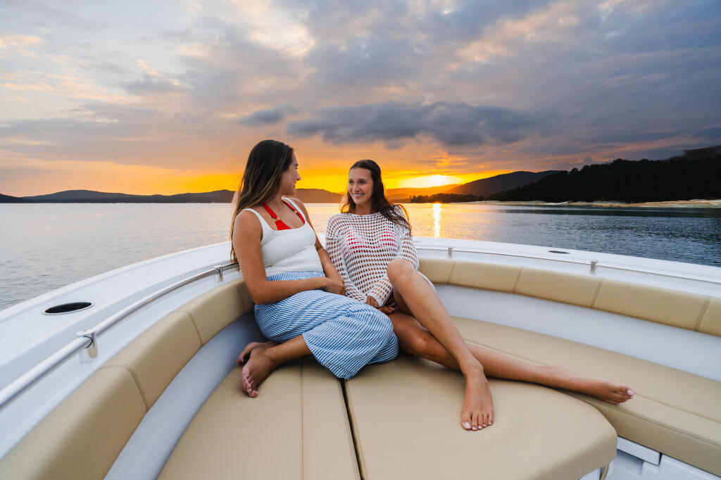 Two women sitting on cushioned bow seating, enjoying a sunset over calm lake waters