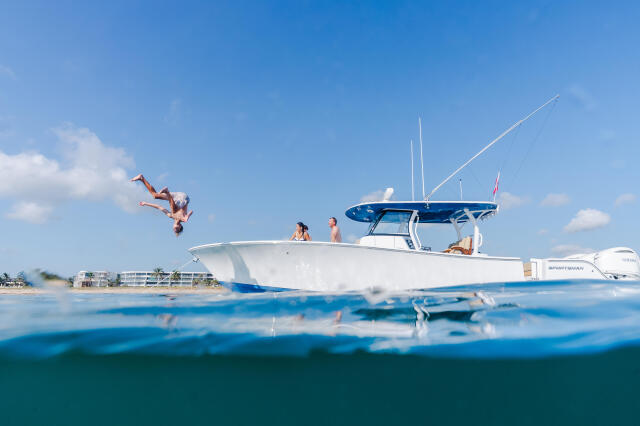 Person jumping off a white and blue sportfishing boat near a sandy beach with other passengers aboard