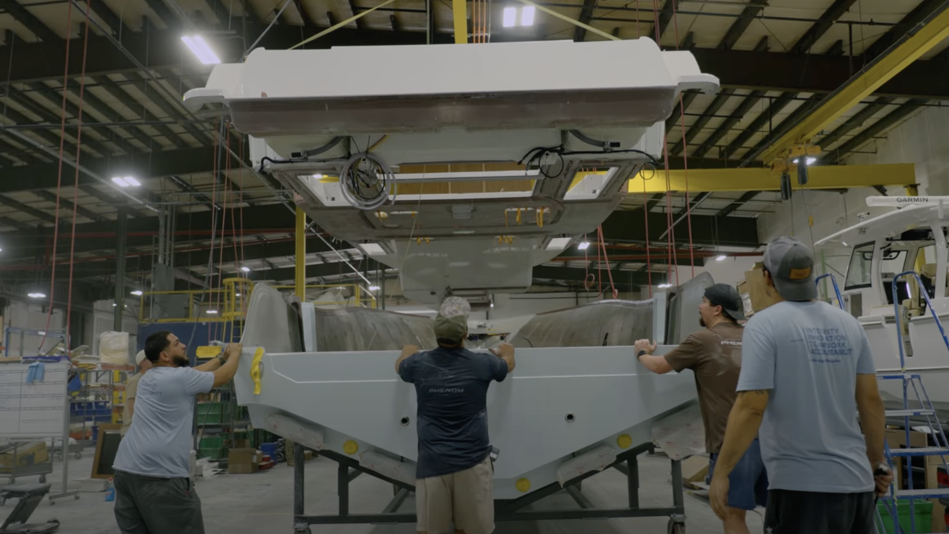 Boat manufacturing workers installing a white fiberglass cabin or hard-top structure onto a hull in a factory facility