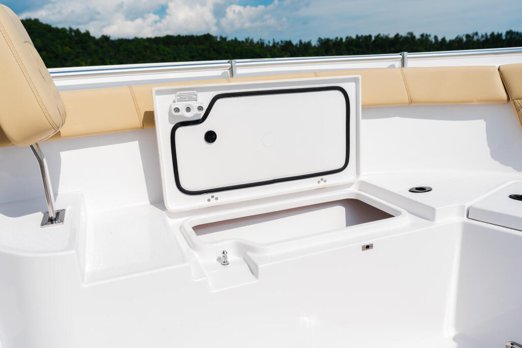 Open livewell with white interior and black lid on boat deck with tan seats and forest backdrop