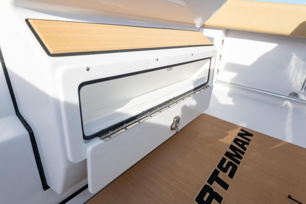 Open storage compartment with tan upholstered lid and white interior panel on boat helm area