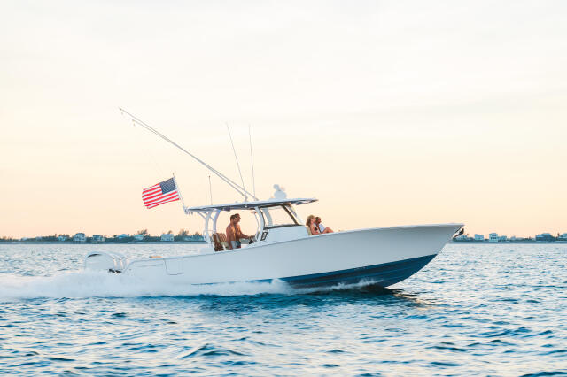 Couple cruising in a white center console boat with hard-top and fishing rods, American flag flying, coastal waterfront in background