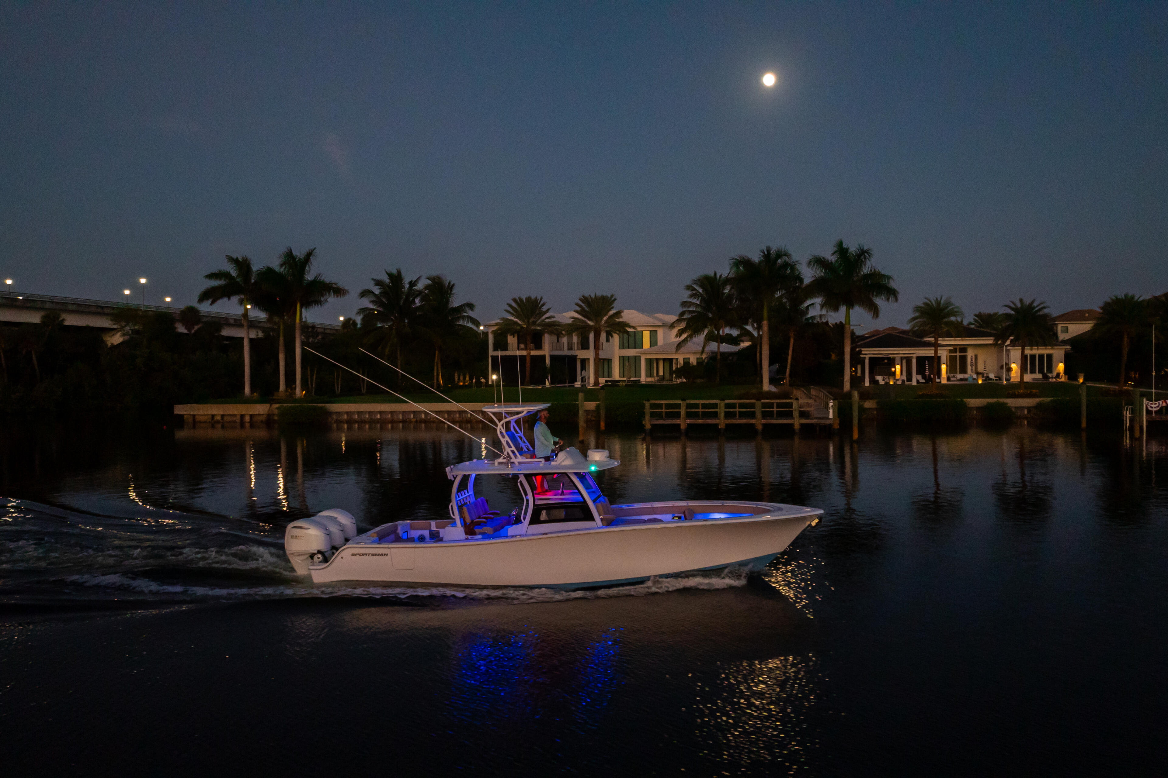 Illuminated center console boat docked at a moonlit marina with waterfront homes in background
