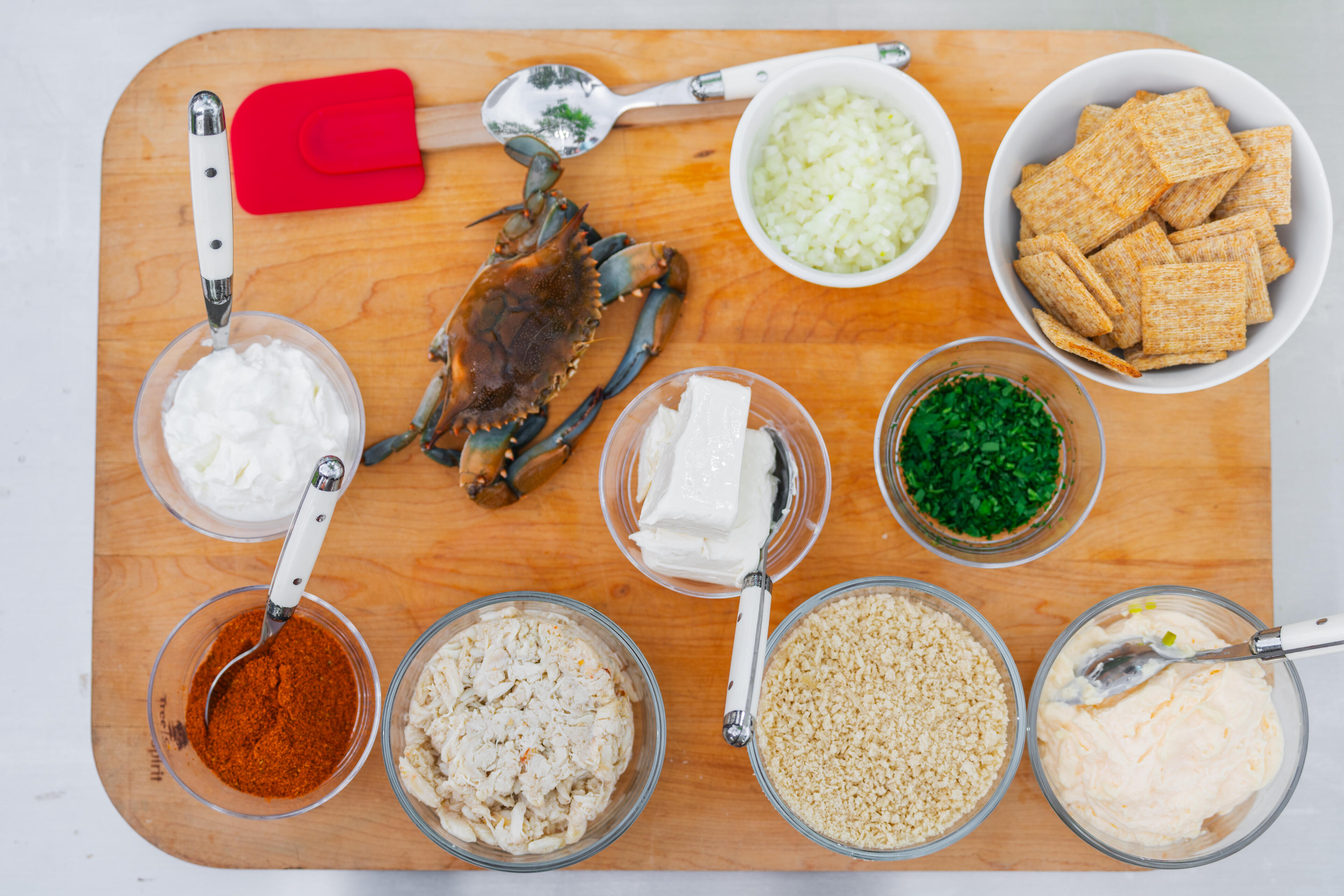 Overhead view of crab cake ingredients arranged on a wooden cutting board including whole crab, breadcrumbs, mayo, spices, and fresh herbs