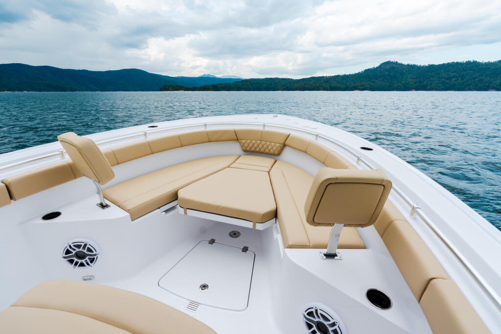 Bow seating area of a white boat with tan upholstered cushions overlooking a forested lake with mountains