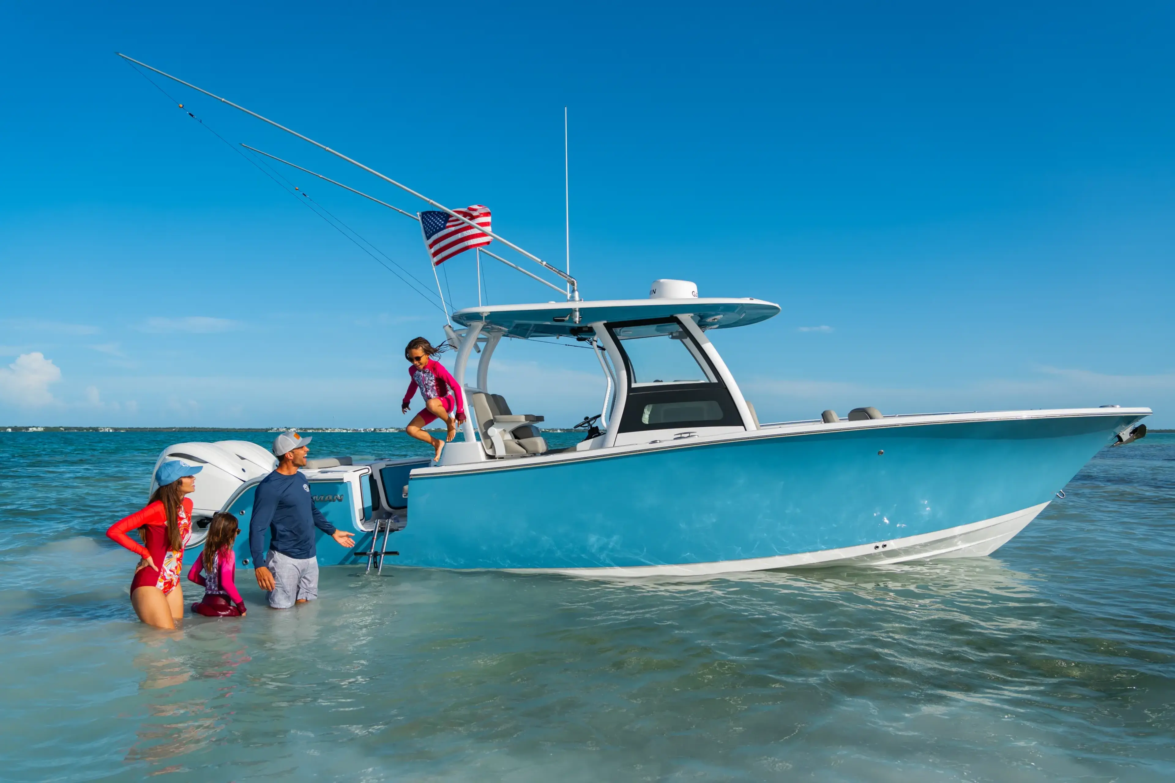 Family wading in clear shallow water next to a turquoise and white boat with fishing rods and American flag