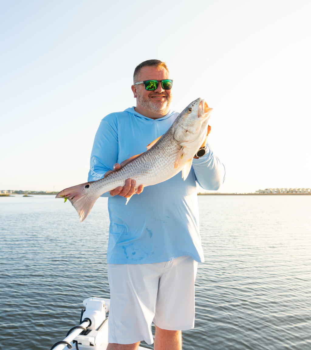 Angler in blue shirt holding a large redfish while standing on a boat in shallow coastal waters