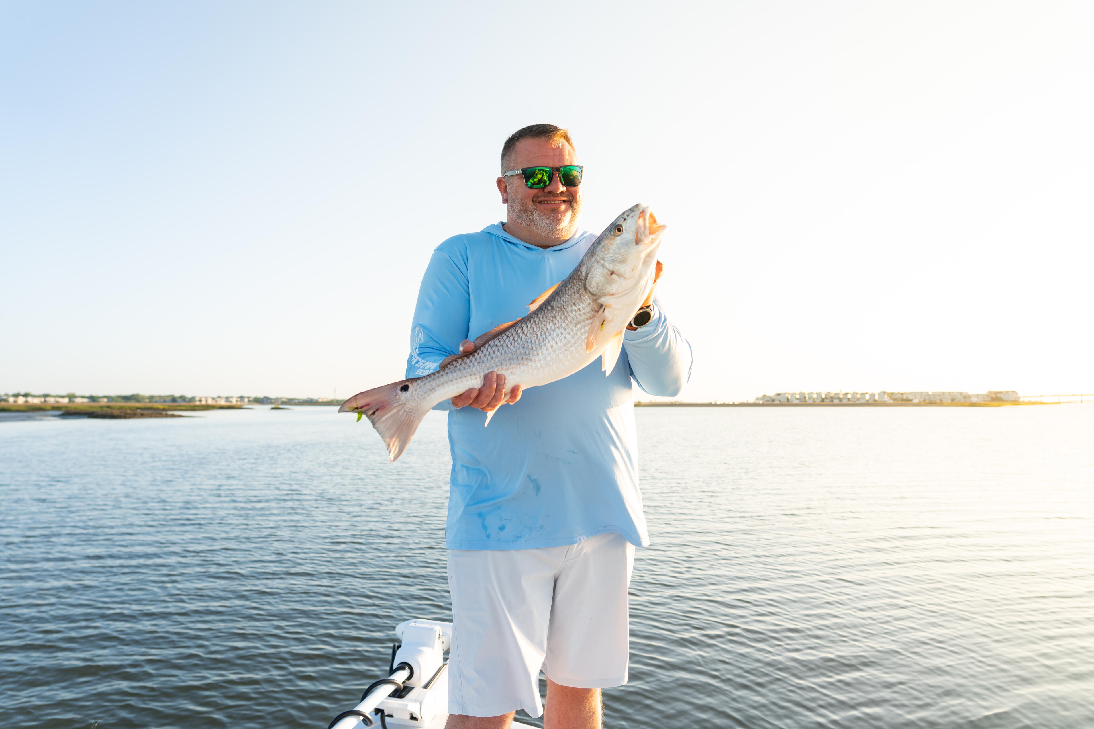 Angler in blue shirt holding a large redfish while standing on a boat in shallow coastal waters