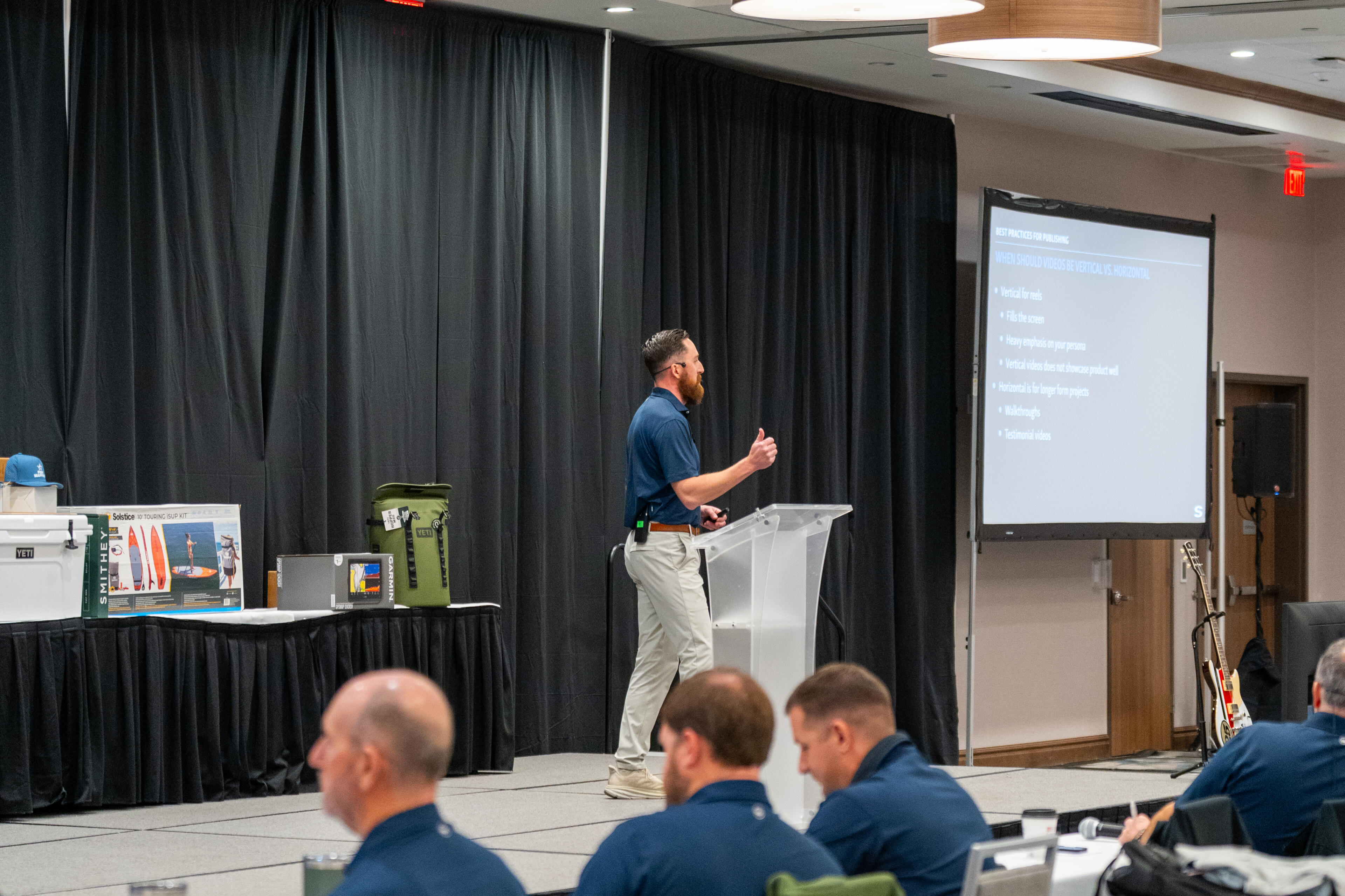 Sales trainer presenting to seated audience in modern conference room with projection screen and wood-beam ceiling