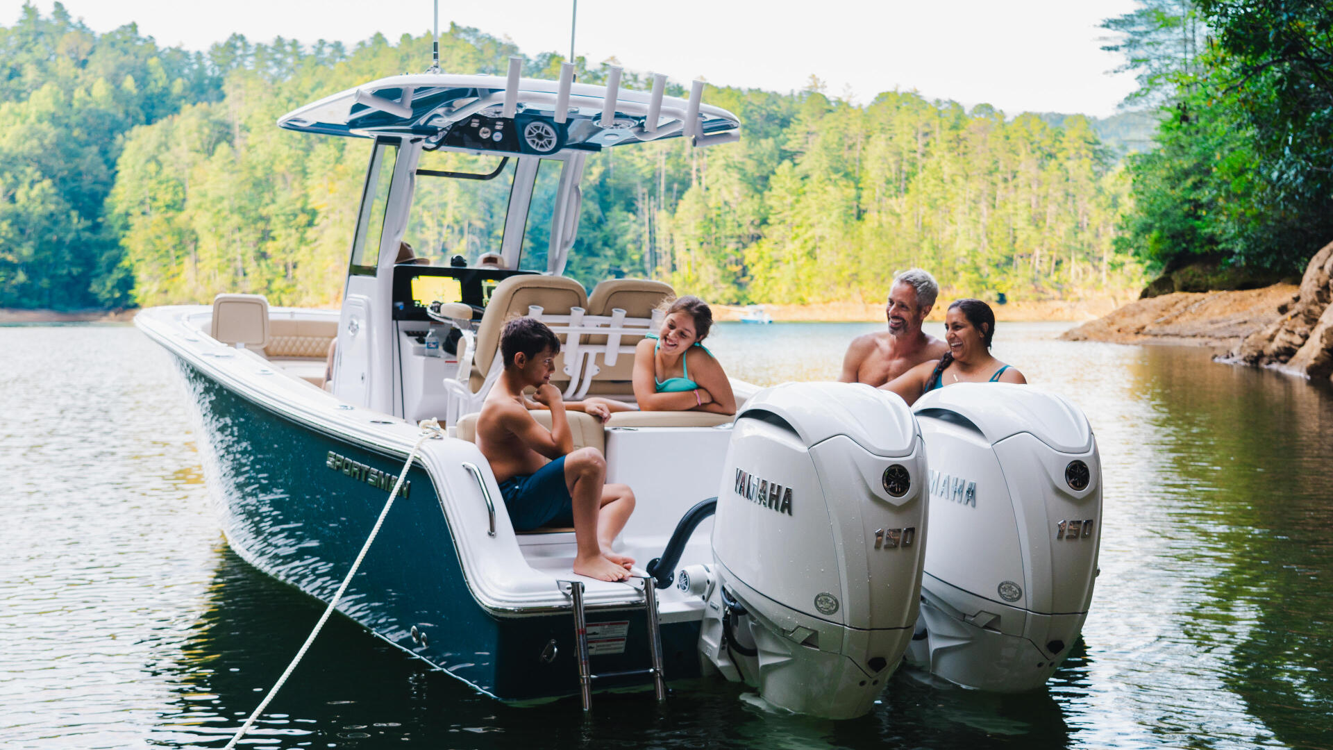 Family relaxing on a dual-engine boat anchored in a scenic lake cove surrounded by forested shoreline