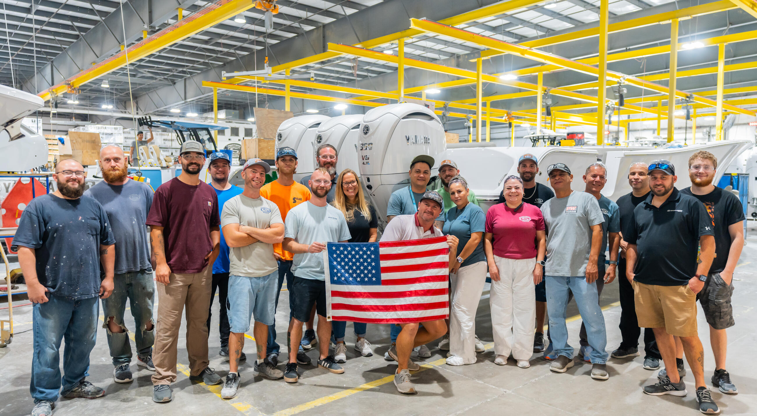 Factory team holding American flag in manufacturing facility with boat hulls and yellow overhead cranes