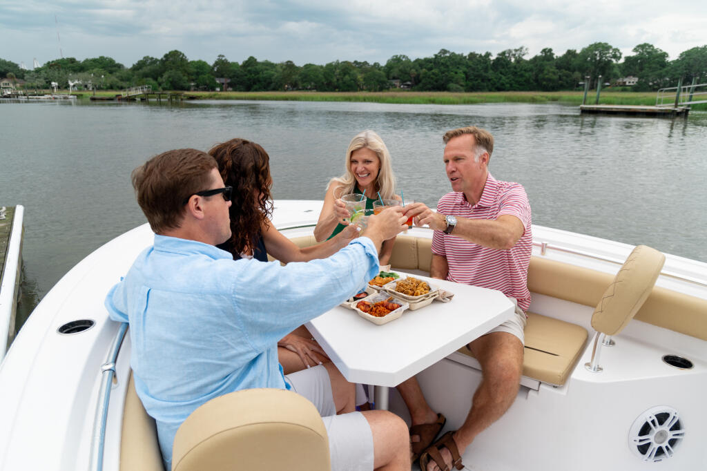 Four friends toasting drinks while dining at a table on a boat in a calm river