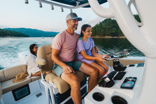 Father and two children at boat helm with watersports equipment on a lake surrounded by forested shoreline