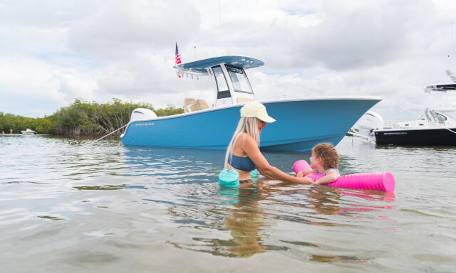 Woman and child swimming in shallow water near a blue center console boat anchored at a sandbar