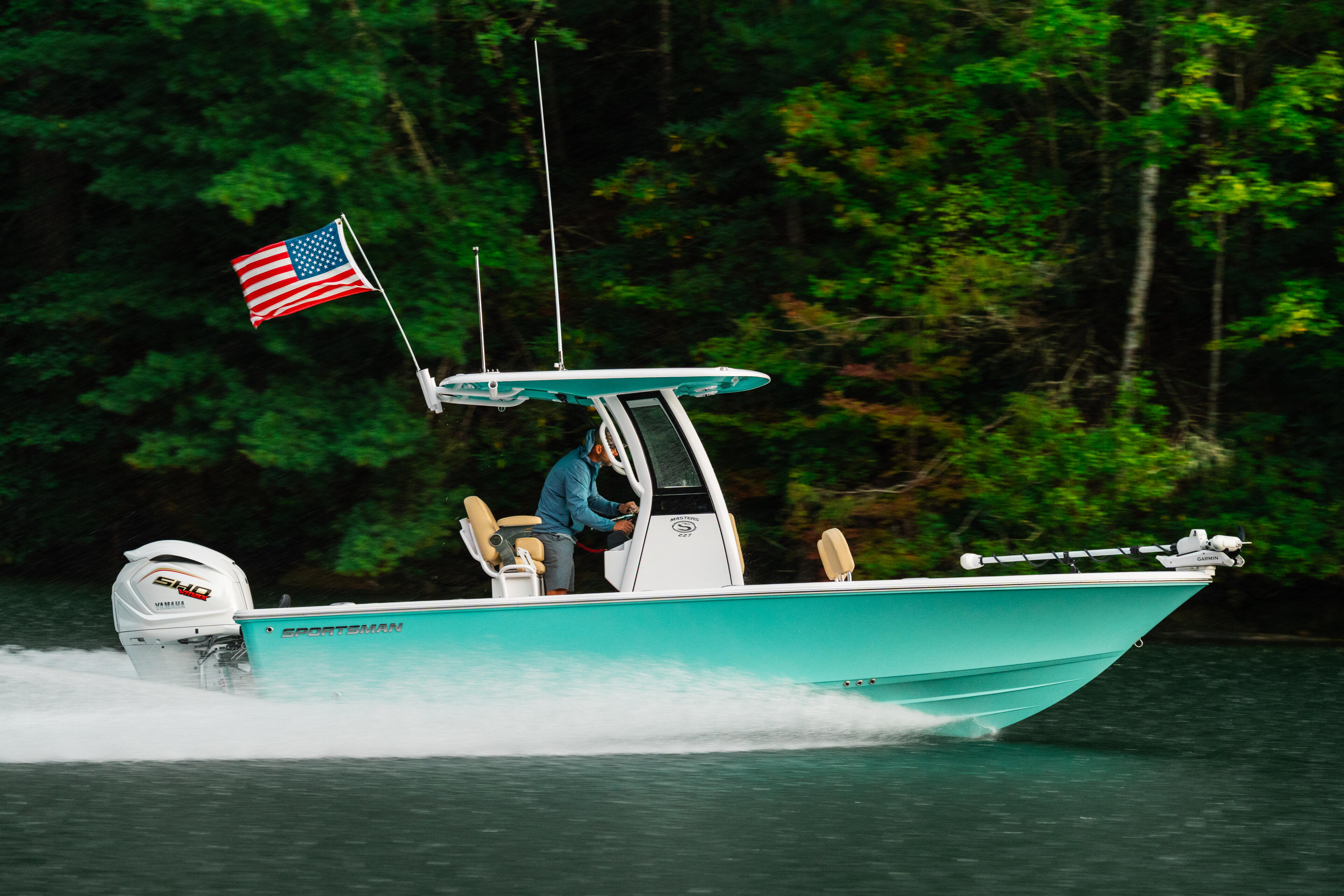 Turquoise boat with American flag running at speed on calm green water backed by dense forest