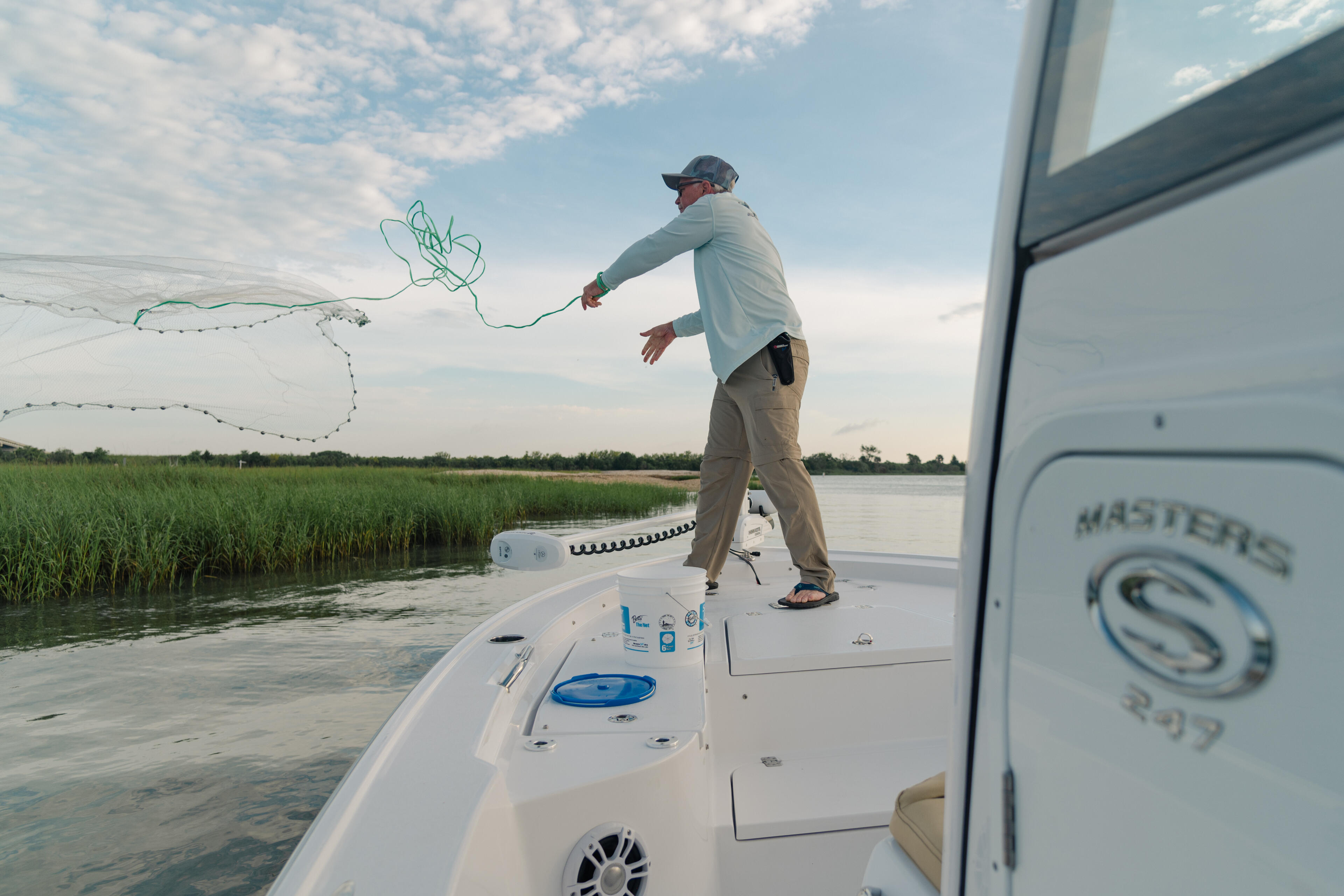 Angler casting a net from the bow of a Masters boat in shallow coastal waters with marsh grass visible