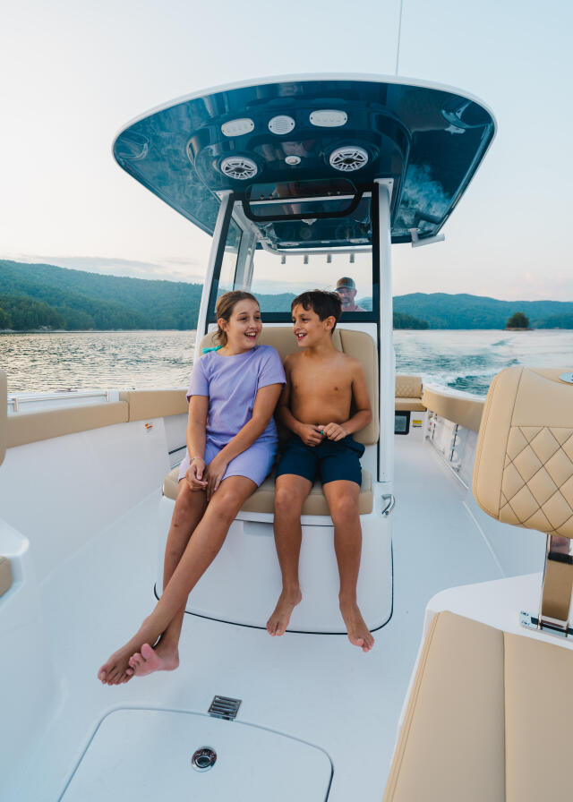 Two kids sitting together on a boat's seating area with a blue hard-top canopy and mountainous lake views behind them