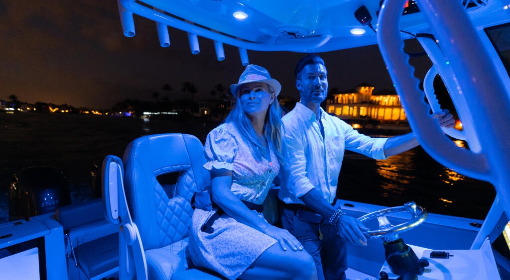 Couple at the helm of a center console boat at night with blue ambient lighting and waterfront lights in background