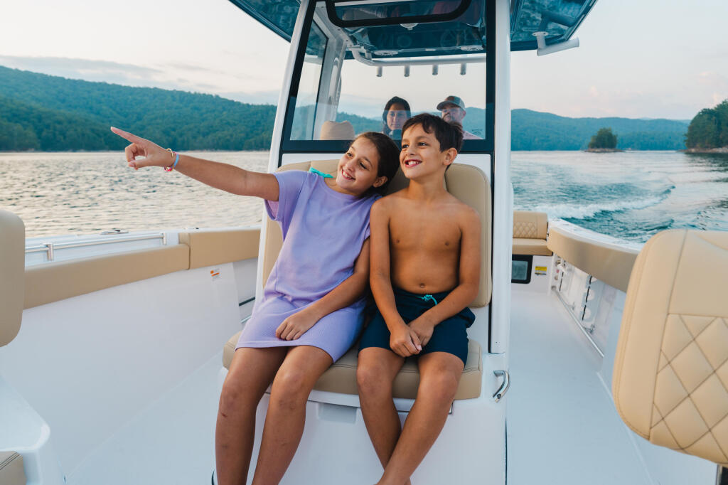 Children seated in the helm area of a boat pointing across a scenic mountain lake