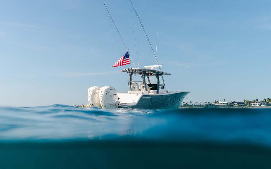 White and teal center console boat with twin outboard engines and American flag anchored in shallow turquoise water near a palm-lined shore