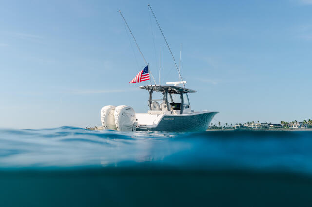 White and teal center console boat with twin outboard engines and American flag anchored in shallow turquoise water near a palm-lined shore