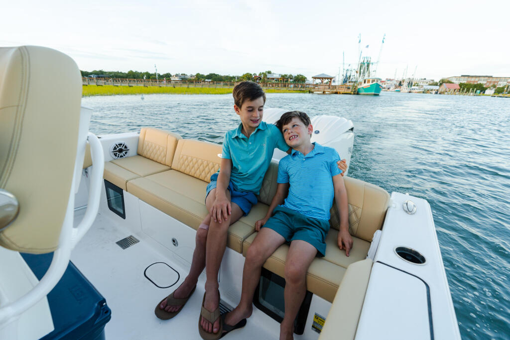Two boys sitting together on cream-colored seating inside a boat on the water near a marina