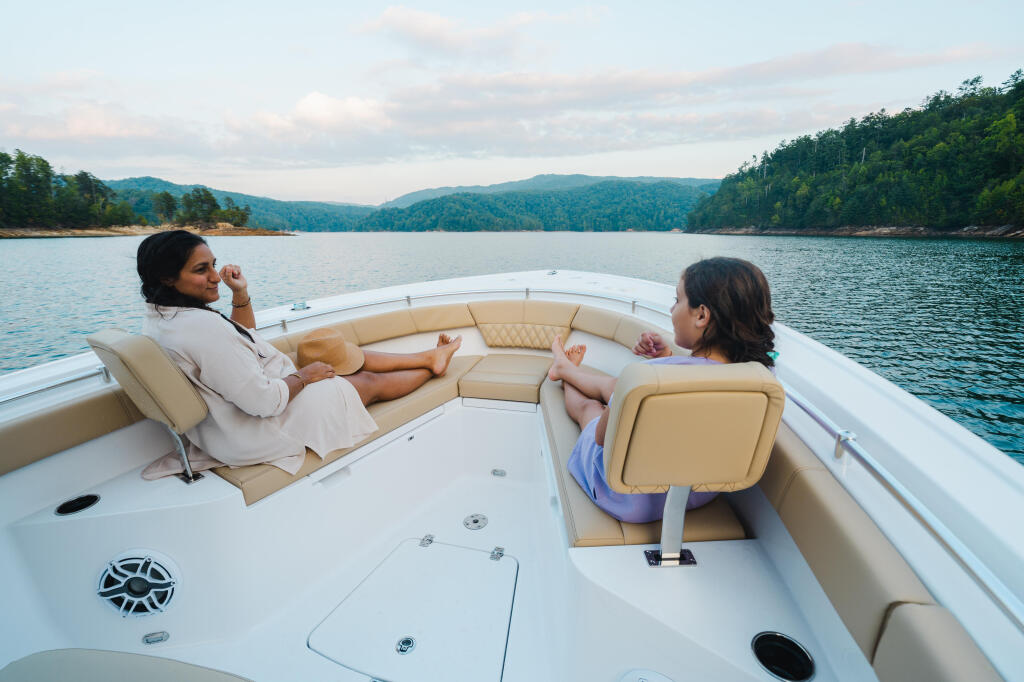 Two people relaxing on comfortable tan seating in a center console boat on a calm lake surrounded by forested mountains