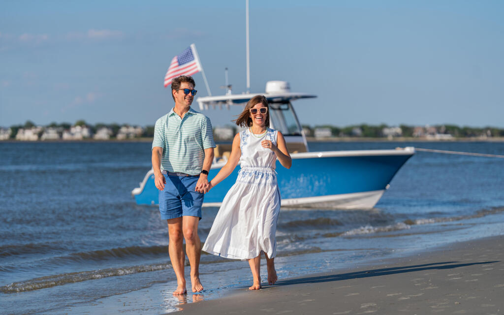 Couple jogging along the beach with an American flag and blue sport fishing boat anchored in shallow water behind them
