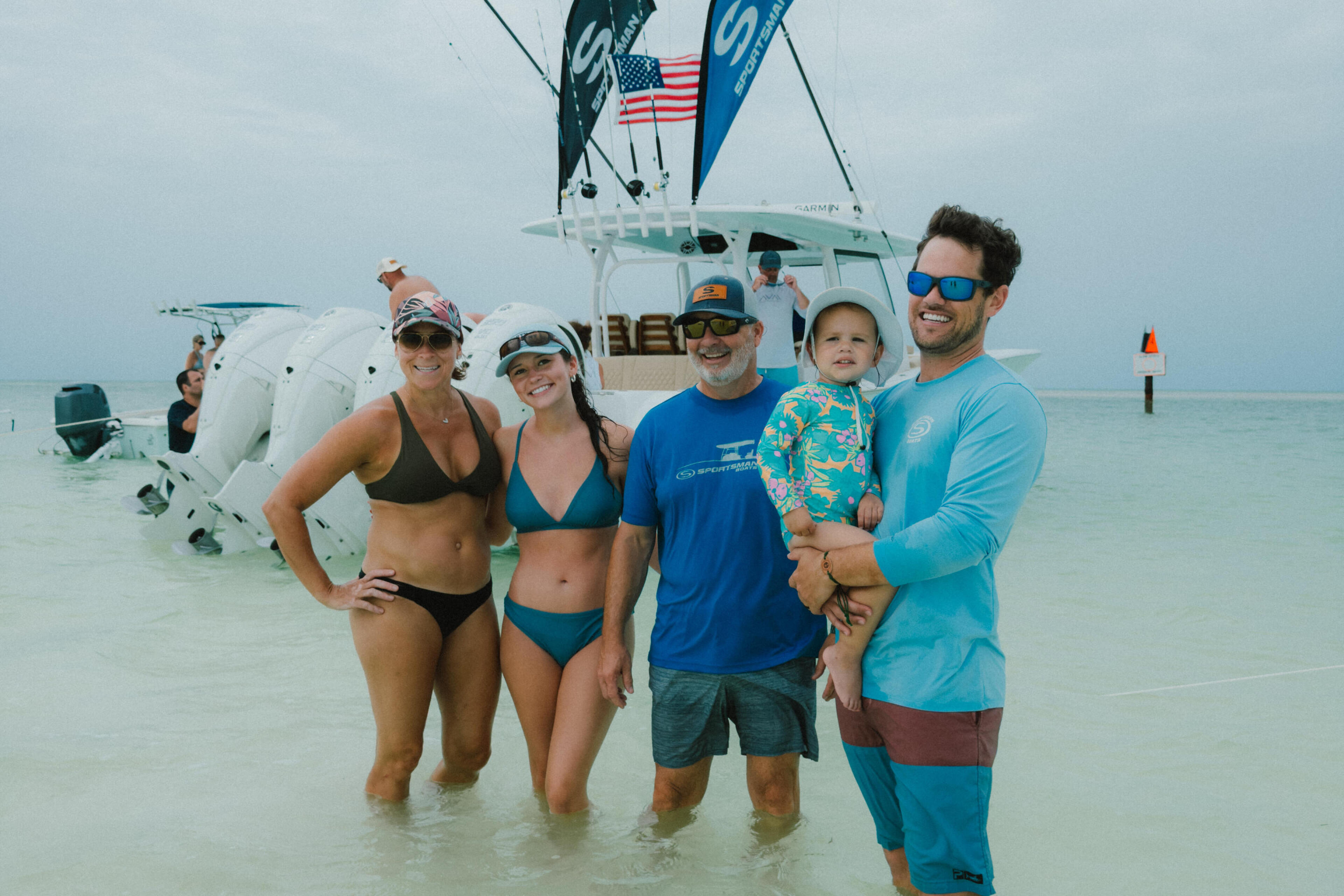 Family and friends pose in shallow water with a white boat displaying colorful kites in the background