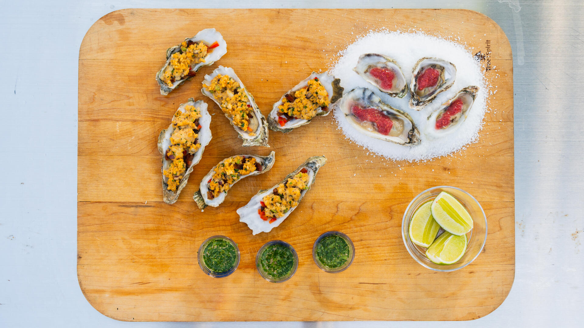 Wooden serving board with prepared oysters, topped with sauce and breadcrumbs, alongside fresh oysters in sea salt and lime wedges