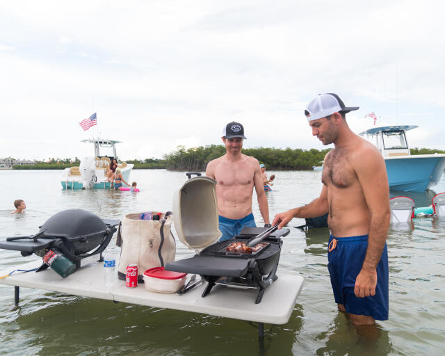 Men grilling food on a floating portable grill at a sandbar gathering with boats and swimmers nearby