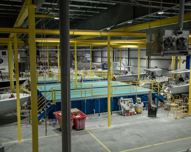 Boat manufacturing facility with yellow overhead cranes and blue platform structures, engines and marine components visible throughout the industrial floor