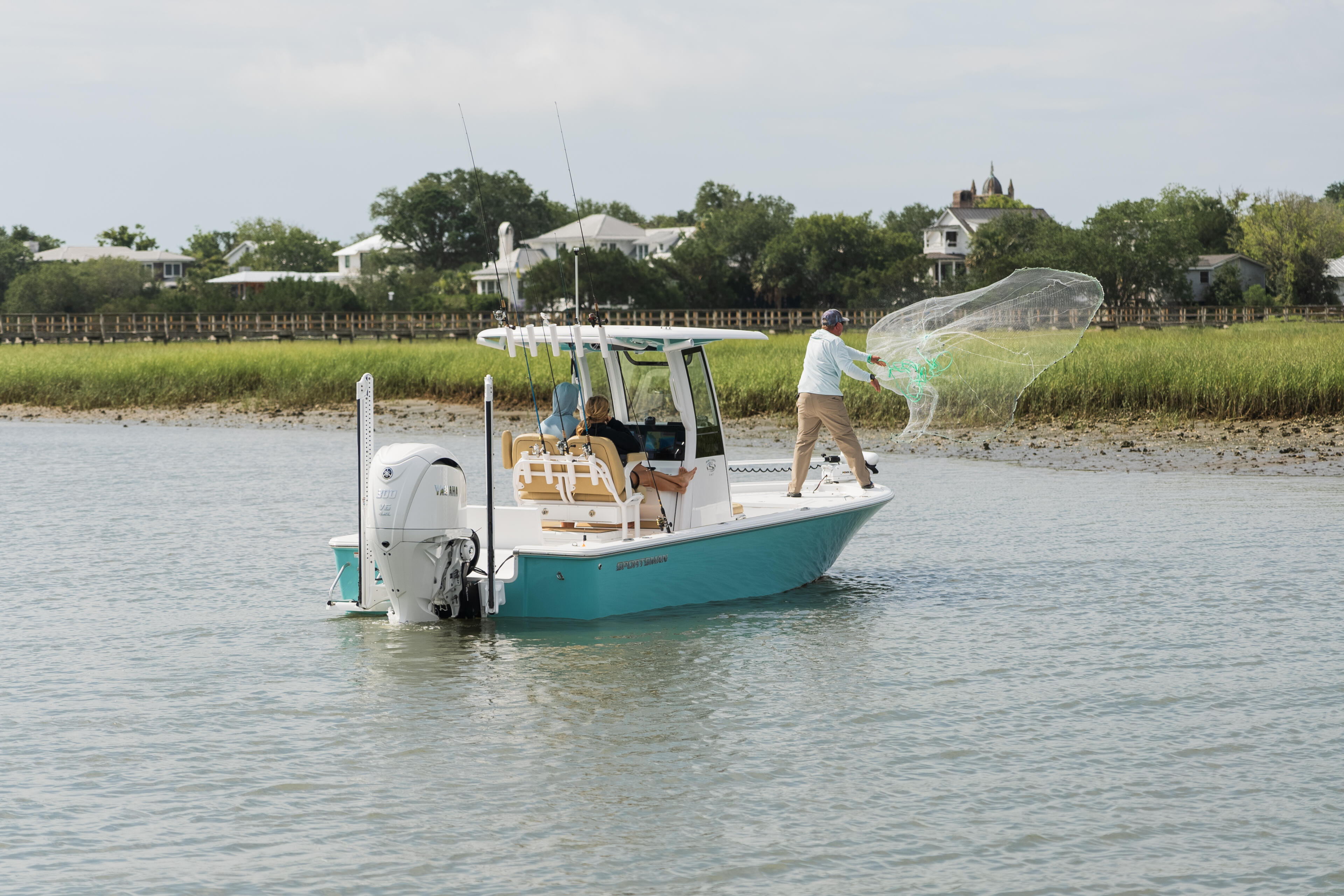 Man casting a fishing net from a turquoise and white center console boat in shallow coastal waters near a marsh shoreline