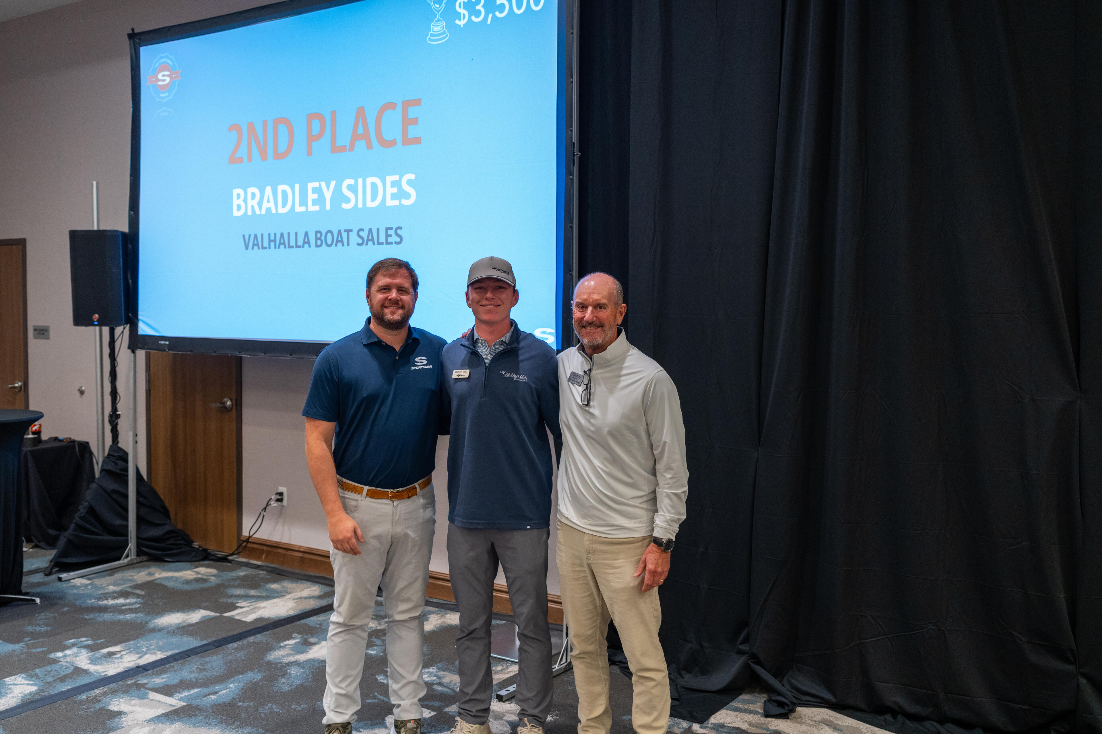 Three men stand in front of a projection screen displaying 2nd place award for Bradley Sides at Valhalla Boat Sales