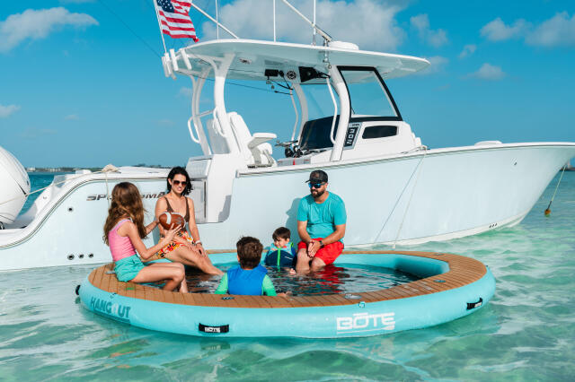Family of five enjoying a floating platform in shallow turquoise water with a sportfish boat anchored behind them