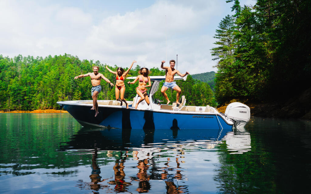 Group of friends jumping off blue boat into calm lake surrounded by forested shoreline