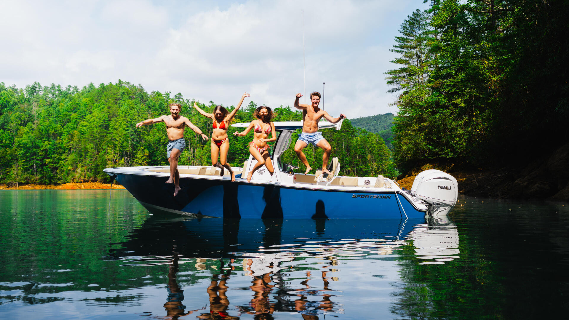 Group of friends jumping off blue boat into calm lake surrounded by forested shoreline