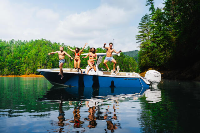 Group of friends jumping off blue boat into calm lake surrounded by forested shoreline