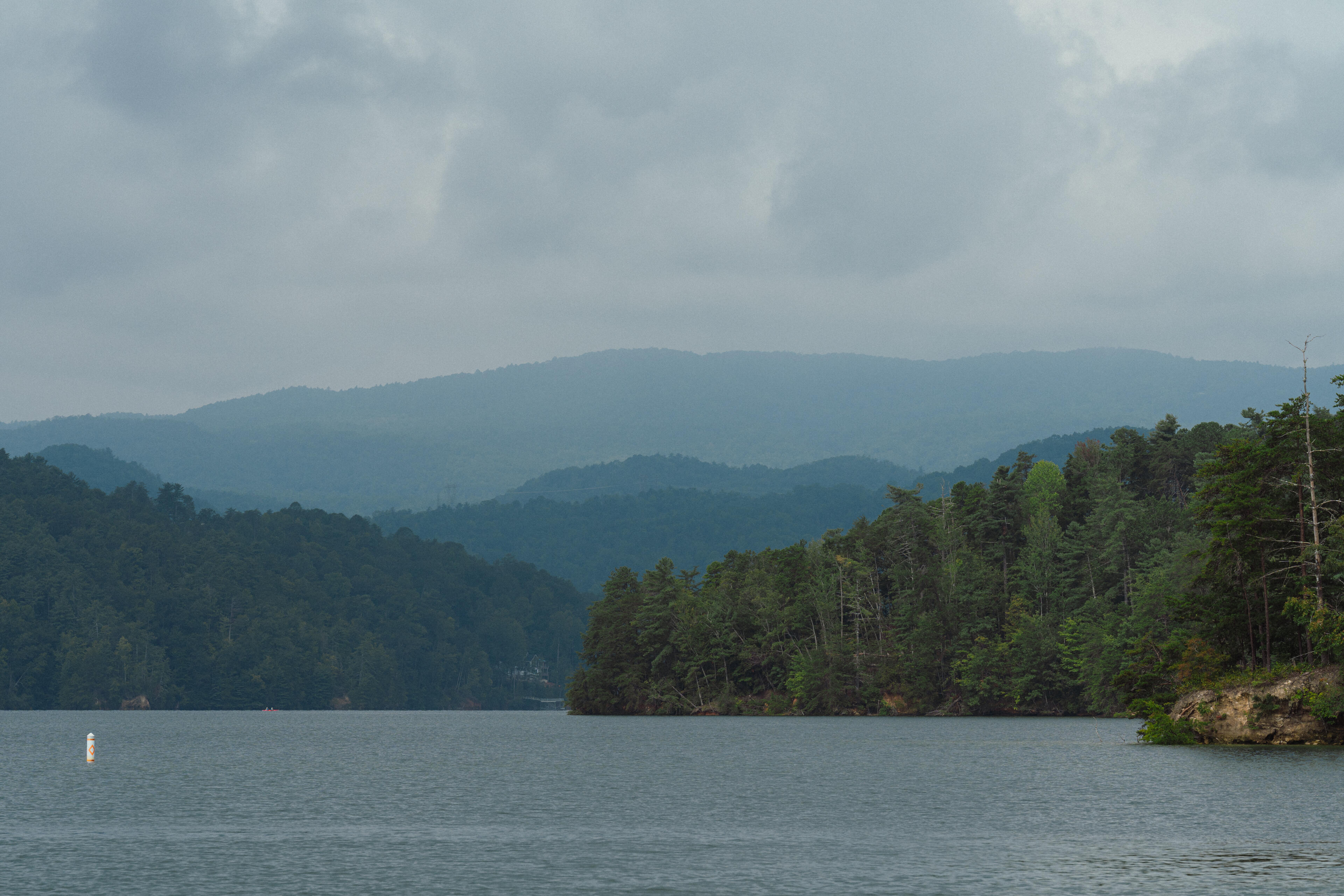Forested mountains rise above calm lake waters under overcast skies with a navigation buoy in the distance