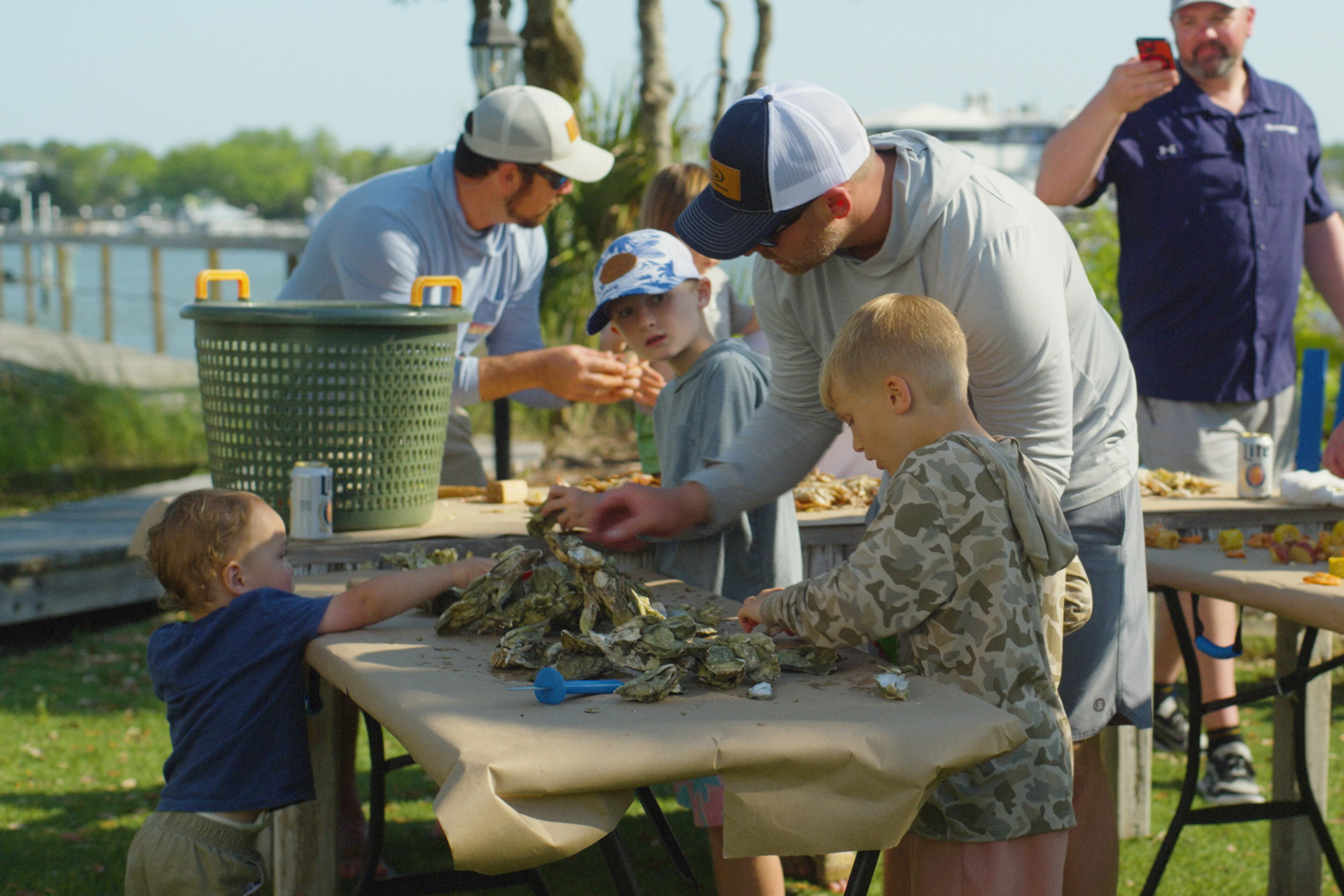 Adults teaching children how to pick and handle fresh oysters at a waterfront marina gathering