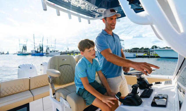 Adult and child at the helm of a white boat with a marina visible in the background