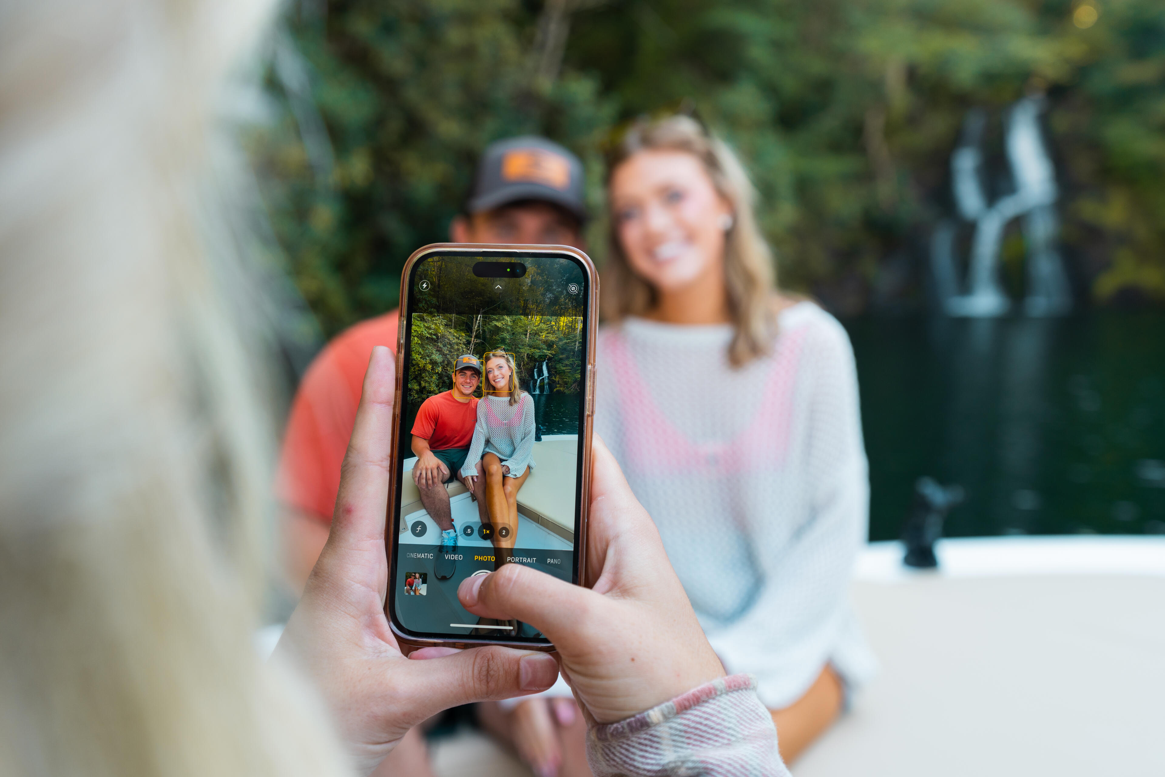 Person holding up smartphone to photograph a couple sitting together by a sandbar with forested background