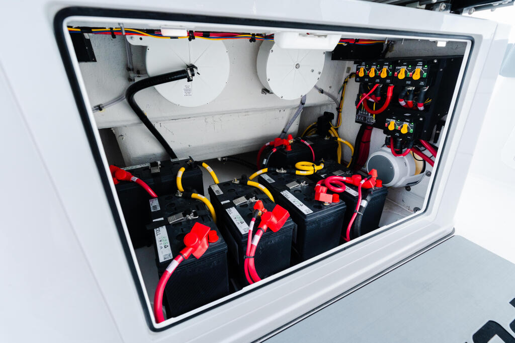Boat engine compartment showing dual batteries with red cables, white fuel tanks, and electrical control panel with yellow switches