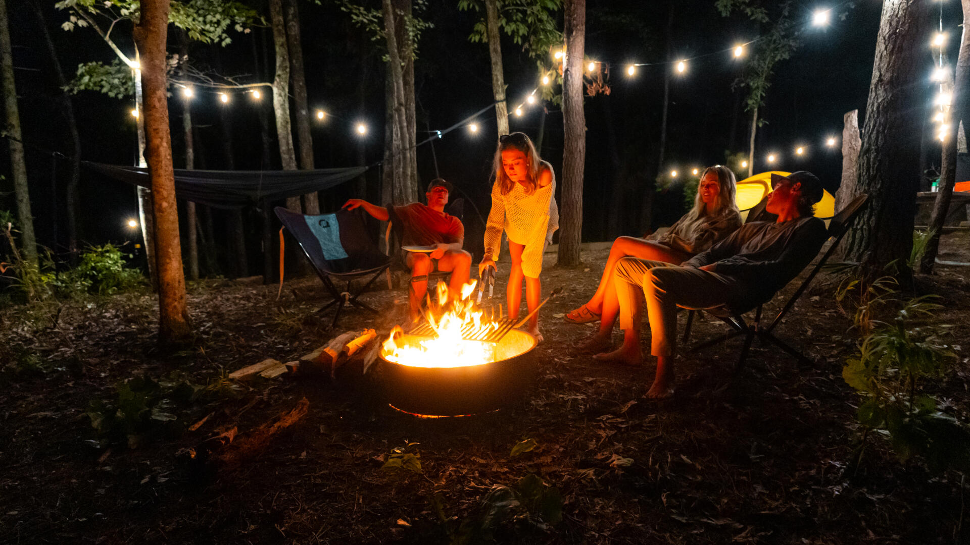 Group gathered around a campfire at night under string lights among tall trees