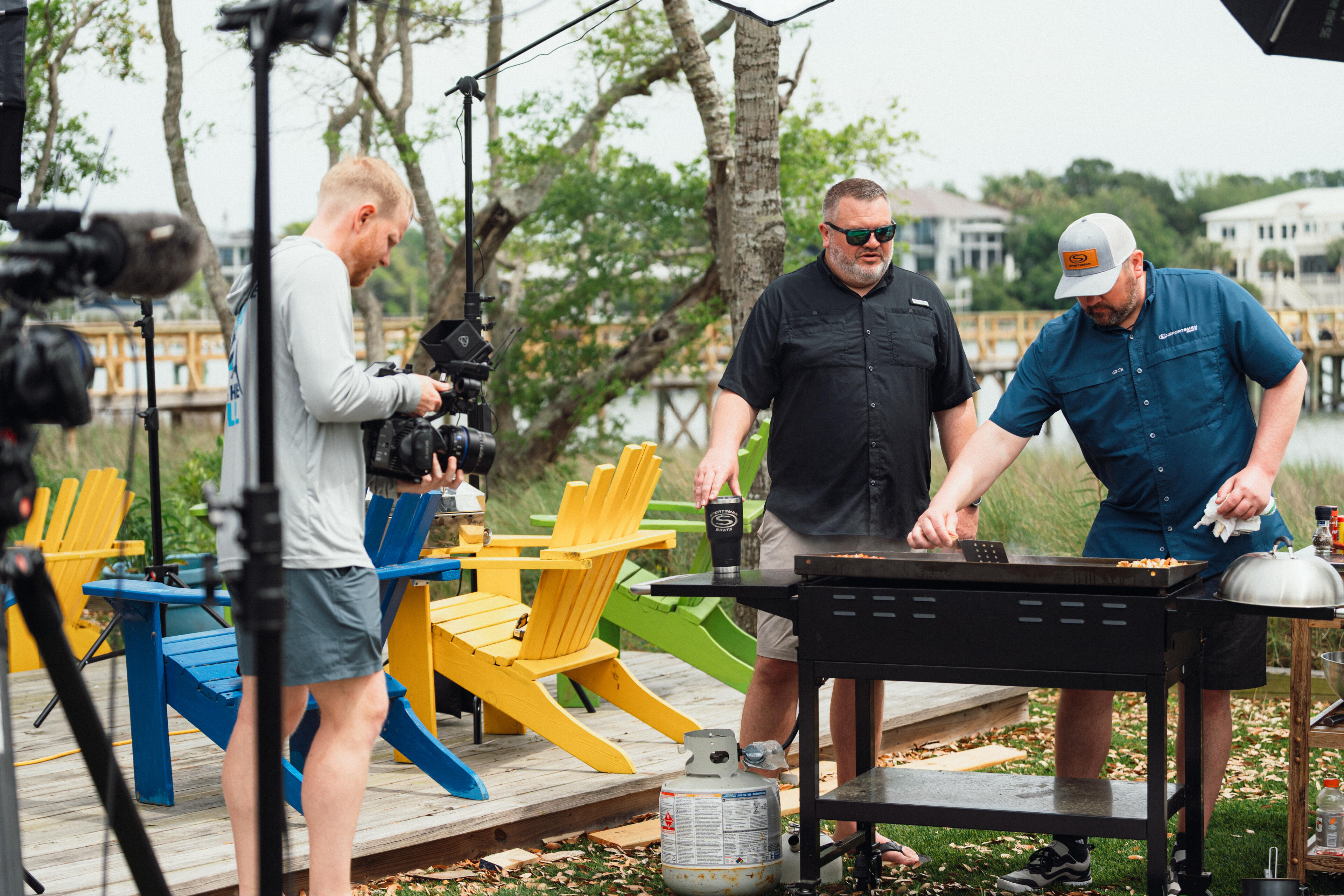 Cameraman filming two men grilling food at a waterfront deck with colorful Adirondack chairs and marina homes in background