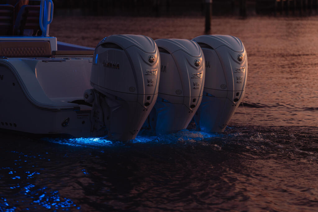 Three outboard engines mounted on the transom of a boat at sunset with bioluminescent water glowing blue