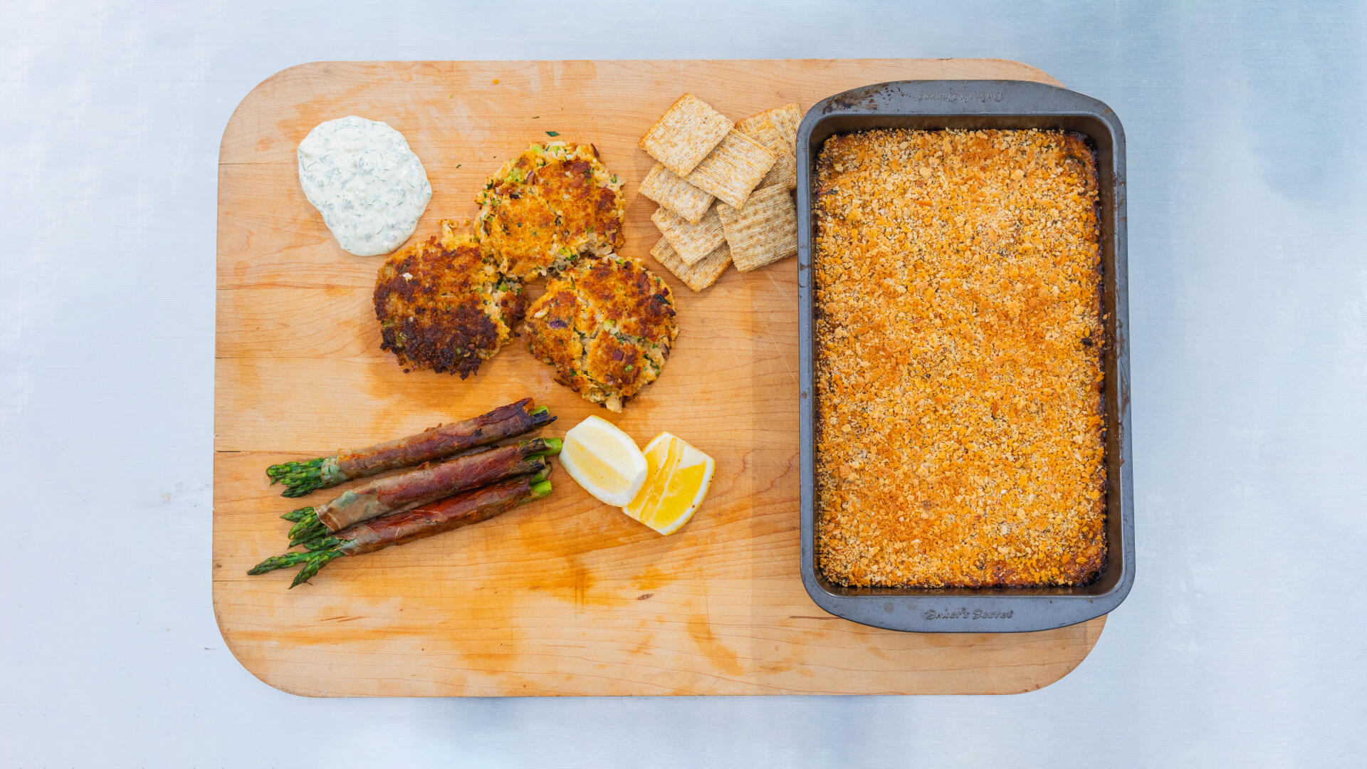 Wooden board with crab cakes, asparagus, lemon butter, crackers, and a baking dish of crab dip against light blue background